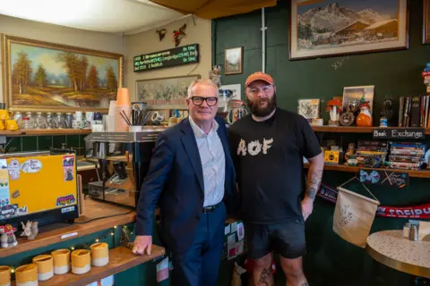 West Midlands Combined Authority Two men, one with short grey hair and black glasses, the other with an orange cap and black beard, are stood next to each other in a coffee shop. 