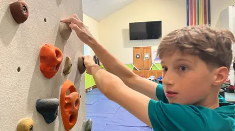 BBC Alex Hutchinson on a climbing wall. He is wearing a blue t shirt and has short brown hair. 