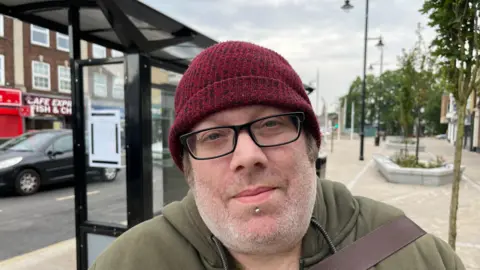 John Devine/BBC Craig Denyer, 43, has stubble on his chin and a dark red bobble hat on his head. He has glasses with dark frames and a gold stud just under his bottom lip. He is wearing a khaki hoodie with the strap of a bag over his shoulder. Behind him is a bus stop with cars passing on the road.