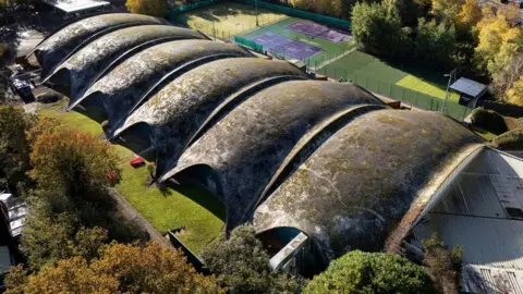 An aerial photo of a row of shell-like concrete structures forming a roof to an oblong building. The concrete is grey and patched with black and yellow lichen. Sports courts and a car park are behind the building.