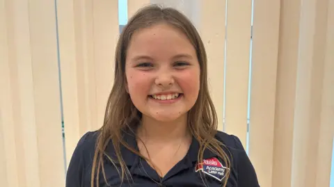 A young girl with long light-brown hair. She is wearing a dark blue and red top with her school's logo on it. She is stood in front of some cream-coloured window blinds.