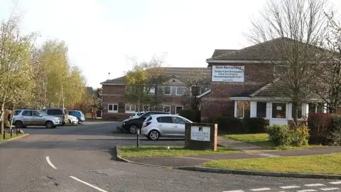 A care home with cars parked in the car park, a sign on a brick wall, and a red brick building, with a sign on it. There is a large white door to the right and trees to the left and right. 