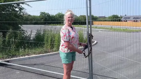 A woman standing in front of a padlocked car park. She has blonde hair and is wearing glasses, a floral top and green shorts.