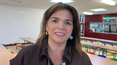Catherine Dourish, a woman with shoulder-length brown hair, stands in a classroom smiling at the camera. She is wearing a brown blouse and colourful earrings. There are wooden desks with multi-coloured seats in the background. 