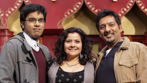 Image of the Masood family from EastEnders. On the left is Tamwar Masood played by Himesh Patel wearing a grey jacket, maroon v neck sweatshirt and a white checked shirt. He has black hair and wears glasses. In the middle is Nina Wadia who plays Zainab Masood wearing a black top and has short black hair. On the left is Nitin Ganatra wearing a beige jacket and a dark grey sweatshirt. He has black hair. They are all smiling. 