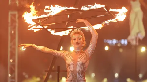 Yui Mok/PA Wire A female acrobat dressed in a gold and white leotard while standing on stage. With one hand, she is spinning a contraption above her head which is lit on fire.