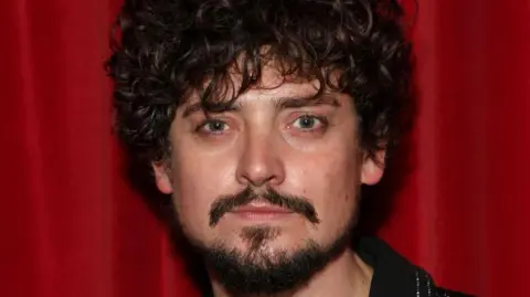 Getty Images A man with curly dark hair and a dar beard and moustache. He is stood in front of a red curtain, wearing a glittery black shirt and looking at the camera with a serious expression.