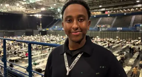 A young man with a black polo shirt and a lanyard saying Nechells stands on a gallery of an exhibition hall with long white tables on the lower level where people sit counting. He has black hair and is smiling.