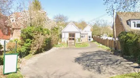 A close-up of the empty church surrounded by houses. The sky is blue with a tree casting a shadow on the driveway in the foreground