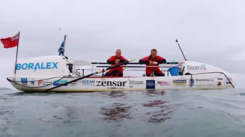 RNLI/Stormy and Steth: The Long Row Home Rowers Mhairi Ross and Allan Lipp posing on their boat while at sea