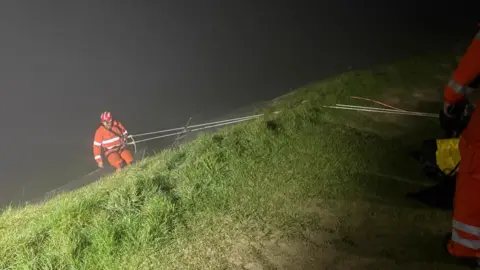 Wiltshire Search and Rescue A man in an orange suit and a red helmet descends a grassy hillside in darkness