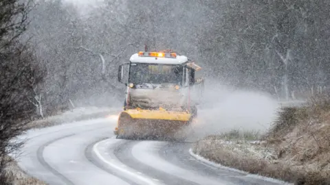 Michael Traill A snow plough travels round a bend on a single carriageway in the countryside, with snow swirling in the air and a thin layer lying on the ground.