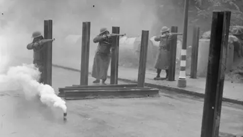 West Glamorgan Archive Service Black and white photograph of the three soldiers from the Home Guard. They are firing rifles with a smoke bomb in the foreground. 