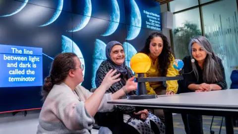 Four people are in a classroom. Three are sat at a table, one is crouched down next to them. On the table are models of the solar system. There is a screen behind them with images of the moon on.