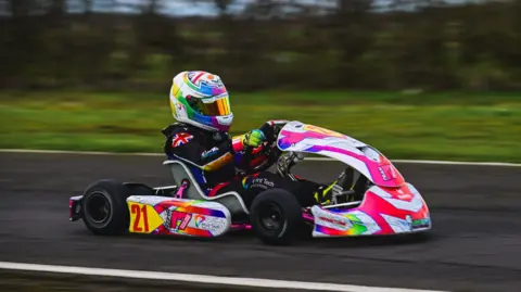 Josh East Photography Deno wearing a colourful helmet with a holographic visor, black racing overalls and bright gloves. He is driving a bright pink and white kart along a black race course. The trees and grass in the background are blurred. 