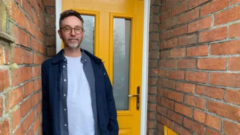 A man is standing in front of his door. It is painted yellow. There is brickwork to the left and right.