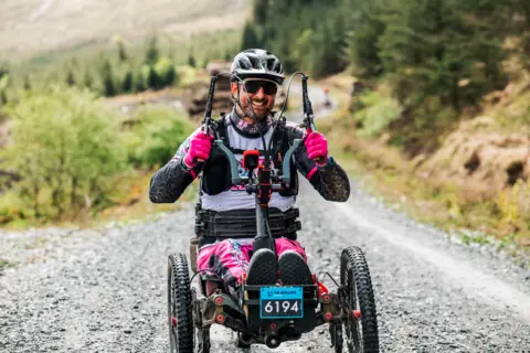 Red On Sports A rider on a recliner bike wearing pink and black gear and a black helmet and sunglasses speeds across the gravel in Galloway with trees in the background