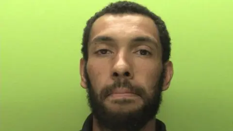 A police mugshot showing a man with short black hair, beard and moustache looking at the camera in front of a light green background.