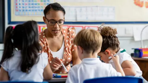 Getty Images Primary school students sitting in a classroom being taught by a teacher. The pupils are learning on mini whiteboards.