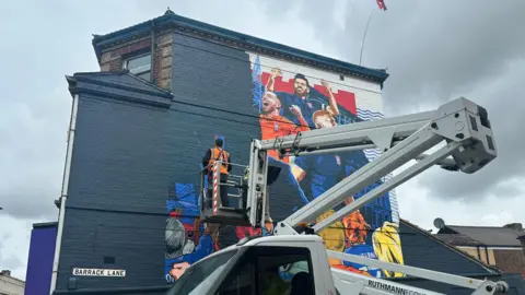 Alice Cunningham/BBC Another angle of the painter on a platform lift paints over a mural that depicted Ipswich Town's players and manager on the side of a building.