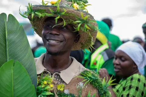 Anthony Siame/EPA/Shutterstock A smiling man wears a straw hat and holds big green leaves - his clothes and hat are adorned with leaves and yellow flowers during a campaign rally in Dar es Salaam, Tanzania - Thursday 28 August 2025