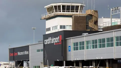 The terminal building at Cardiff Airport, with grey and black walls. The words 'Croeso i Gymru' amd 'Welcome to Wales' can be seen against a black background, while 'Cardiff Airport' is also visible on another wall. An observation tower with windows is in the centre of the frame, with an exterior concrete spiral staircase attached to the right of the tower.