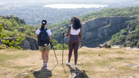 Getty Images Two female hikers - one dressed in gymwear; the other in a t-shirt and shorts - stand on a hillside overlooking a rural town and reservoir. Both have walking poles.