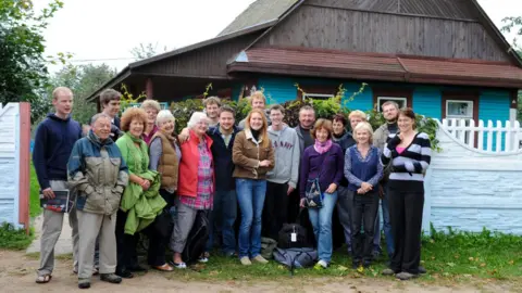 Diana Brooks A group of people wearing jackets, jumpers and scarves stands outdoors in front of a wooden building and a pale blue fence in a rural setting, photographed in 2010.