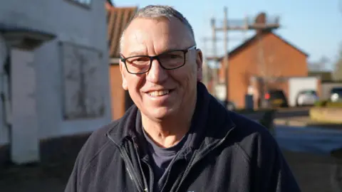 Shaun Whitmore/BBC Sinclair Glover wears a pair of black-framed glasses, a navy T-shirt and a navy fleece. He is standing outside the boarded-up building.