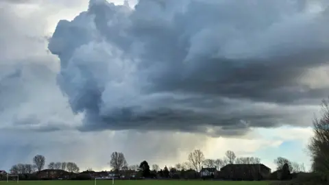 Figaro Large, billowing grey clouds above a grassy field. There are two goalposts and houses and trees cast in shadow in the distance. There is a patch of blue sky peeking through in the right bottom corner.