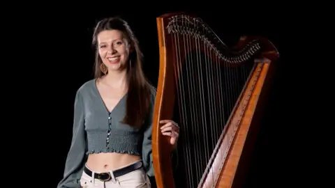 BBC Radio Scotland/Martin Shields Rachel Groves standing with her harp. She has long brown hair and is wearing a green top and white jeans.