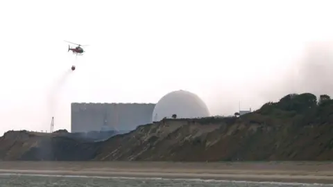 PA Media Un helicóptero transporta agua recogida del mar para arrojarla sobre el incendio forestal en Dunwich Heath, cerca de Leiston, próximo a la central nuclear Sizewell B, donde se ha declarado una emergencia mientras los bomberos luchan contra un gran incendio en los brezales de la costa de Suffolk.