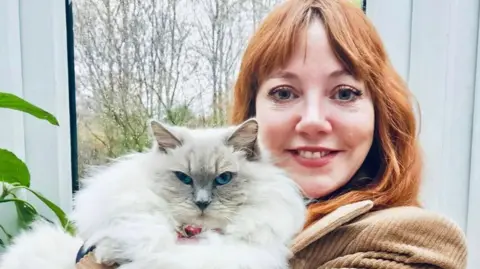 Diane Morgan, pictured holding a large, fluffy white cat which looks directly at the camera. Diane has long red hair and wears a brown corduroy jacket; she is smiling at the camera, standing in front of a window with trees in the background.