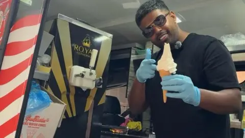 Mr Shuey with a regular ice cream a cone - he has short dark hair, shades, a black T-shirt and blue gloves.