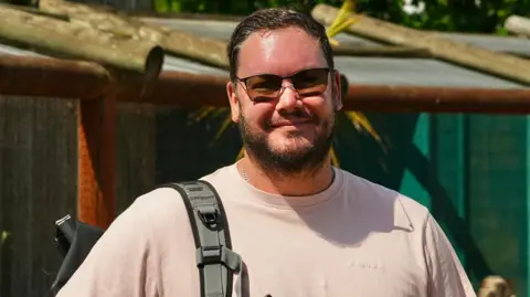 Martin Giles/BBC Graham Hunt smiles at the camera. He has brown hair and beard and wears tinted sunglasses. He has a rucksack slung over his right shoulder. 