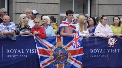 PA Media A crowd of people, many holding Union Flags and Jersey flags, wait behind barriers for the King and Queen.