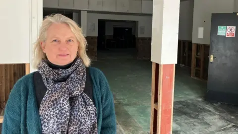 Jo Kitching wearing a scarf and cardigan. She looks at the camera while standing in a stripped out, empty building.