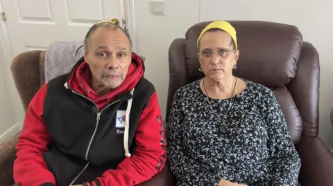 A man with a grey beard is pictured in a red and black hoodie while a woman sits beside him in a white living room wearing a black floral top and yellow headband.