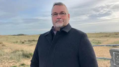 Henry Godfrey-Evans/BBC A man in a dark coat smiling with open grass behind him