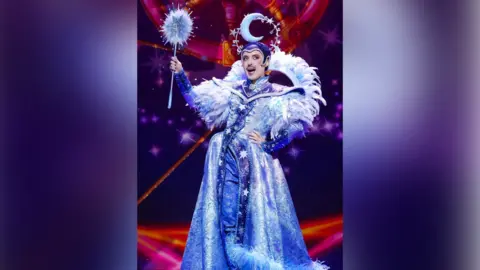 Paul Coltas A person on stage dressed in a purple and blue dress with flowing skirts and purple fur. They have feathers on the shoulders of the dress and are holding a blue wand that is fluffy and star shaped. They have a purple hat with a headdress attached in the shape of a crescent moon