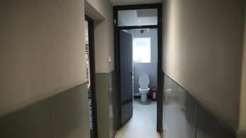 SHAUN WHITMORE/BBC The toilet cubicle is a thin, small toilet in a very narrow corridor off the main clubhouse bar. It shows a while toilet with the lid up and a window above it. It is a small space. The corridor has grey wood panels at the bottom of the wall and cream paint at the top. The floor is lino