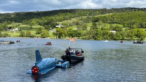 A black boat tows Bluebird out into Coniston Water. A few other boats are dotted around, while tree covered hills rise up on the distant shore.