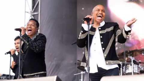 Jackie Jackson and Marlon Jackson singing on a stage. They are wearing black jackets and holding microphones. 