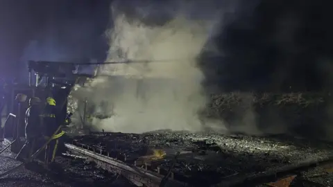 Totnes Fire Station A completely destroyed building which has been burnt down by a fire. There is still smoke from the fire and two fire officers are stood inspecting the scene while holding a fire hose. 