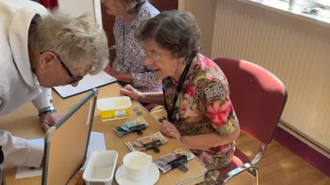 Jennifer is sitting in a chair. She is wearing a leopard print and floral top and is peering over a table. There are bank notes in front of her. Another woman is bending down writing on a piece of paper on the table. 