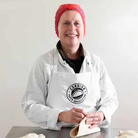 John Hersey Professional baker Jackie Tonkin of the Cornish Pasty Association  is smiling at the camera as she crimps a pasty. She is wearing a white coat, a white apron with a CPA logo and a red hair net. 