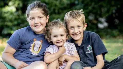 Lucy Lucy's three young children hugging one another and smiling at the camera. They are outside on the grass and wearing tshirts.