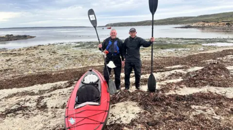 Cockleshell 22 Image shows two men on the shore of a beach standing next to a red, two man kayak. They are holding their oars up next to them. 