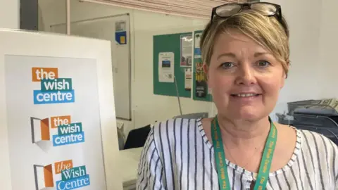 Rebekah Wilson wearing a striped top standing next to a Wish Centre sign in their office building. Rebekah has blonde hair and glasses on her head and is wearing a green lanyard.