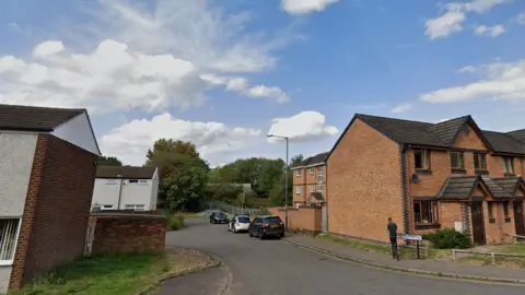 Kingswood Road, Nuneaton, is flanked with red-brick buildings with some cars parked. The road leads on to a grassed area with trees visible